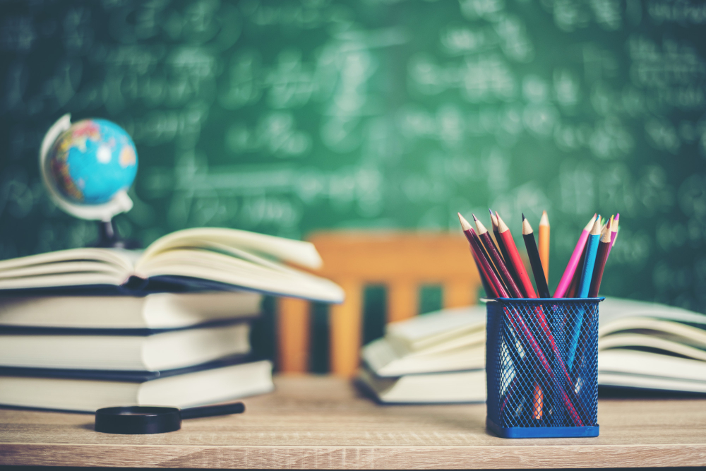 kobi education-esai iisma-gambar tumpukan buku dan pensil di atas meja guru di dalam ruang kelas